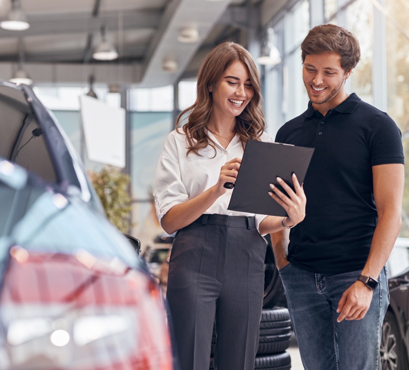 Kobieta trzymająca podkładkę z klipsem, obok mężczyzna uśmiechający się czytający z podkładki, w lewej części kawałek auta